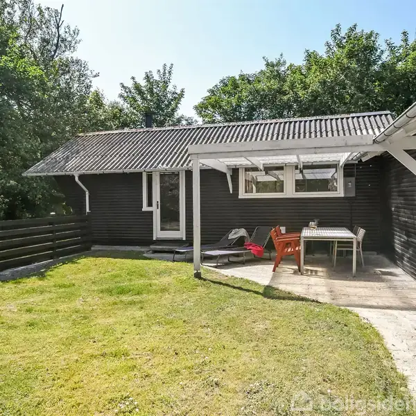 Et sortmalet træhus med en terrasse er udstyret med et bord og stole. Det er omgivet af en grøn græsplæne og træer i baggrunden.