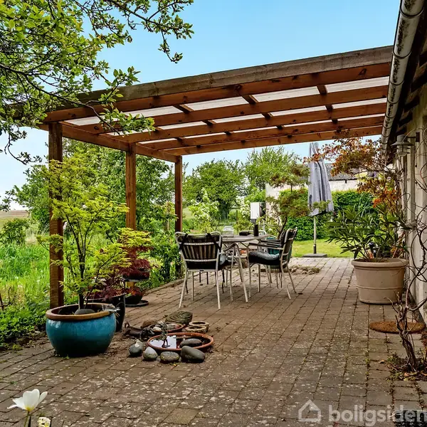 En terrasse med havemøbler under en træpergola omgivet af potteplanter og grønne omgivelser. En stor, flisebelagt plads fører til en frodig have fyldt med træer og buske.