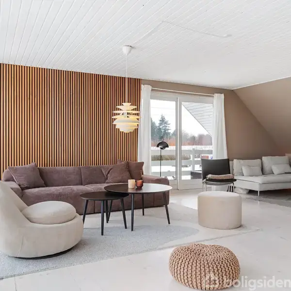 Stue med moderne møblering bestående af en brun sofa, lænestol, og sofabord på gulvtæppe. Bagvæg i træpaneler, glasdør til terrasse med udsigt til sneklædt landskab. Lampe hænger fra loftet.