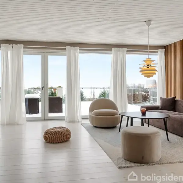 En stue med en stor brun sofa, sofabord og moderne loftslampe. To store vinduer med hvide gardiner viser en terrasse udenfor. Rummet har lyse trægulve og minimalistisk indretning.