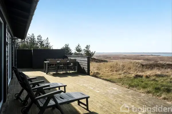 Terrasse med to liggestole og et bord med stole, beliggende ved et sommerhus. I baggrunden ses en mark med græs og buske, der strækker sig ud mod havet under en klar himmel.