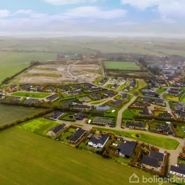 En række beboelseshuse ligger organiseret i et kvarter, omgivet af grønne marker og veje. I baggrunden ses ubebyggede områder og flere trægrupper, der strækker sig mod horisonten.