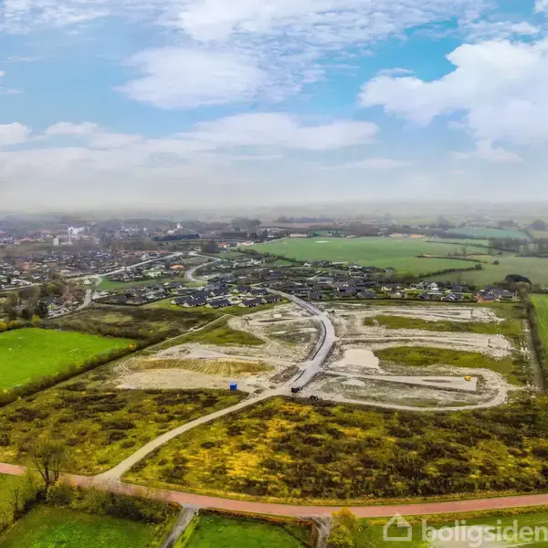 Luftfoto af byggeplads, hvor grunden er delvist klargjort til byggeri. Omkring grunden ses grønne marker og en mindre by i det fjerne.