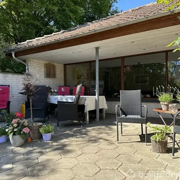 Terrasse med diverse havemøbler og planter i krukker står i solen foran et hus med store vinduer. Bord dækket med dug og stole står under et halvtag.