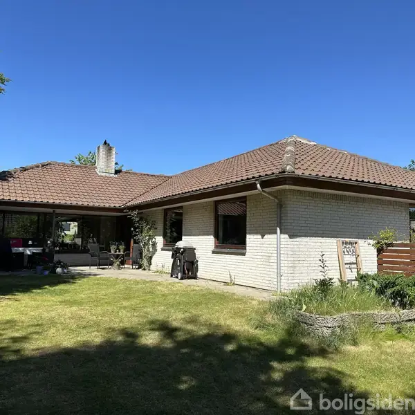 Hvidt murstenshus med brun tegltag, stående i solrig have med græsplæne og planter. Terrasse til venstre med havemøbler, omgivet af træer og blå himmel.