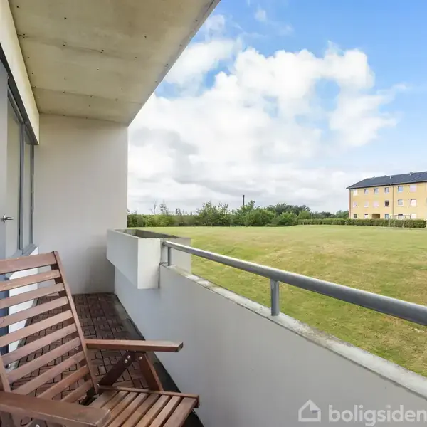 En stue med en grå hjørnesofa står omkring et rundt sofabord. En fladskærms-tv er placeret til højre. Store vinduer viser en udsigt til en boligblok.