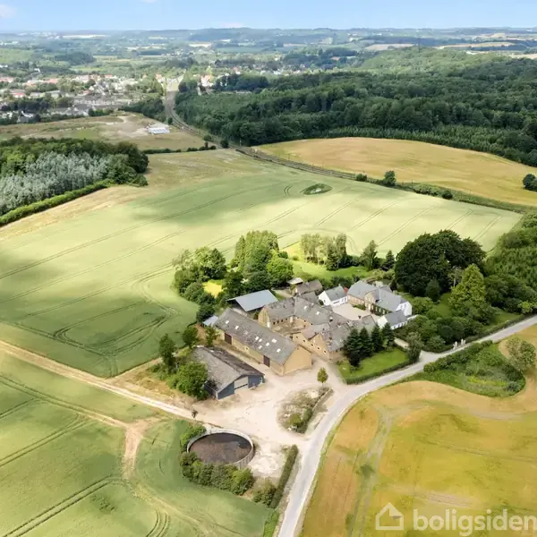 En gård omgivet af marker og skov ses fra oven. Bygningerne er centralt placeret med alléer og veje omkring, beliggende i et landligt landskab.