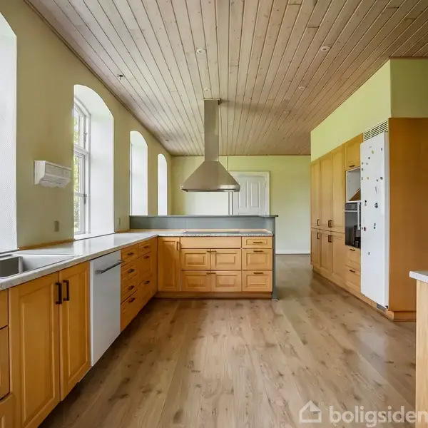 Et rummeligt køkken med lyse træskabe og grå bordplader. Rummet har store vinduer, trægulv og en rustfri stål-emhætte. Baggrunden viser en grøn udsigt udenfor.