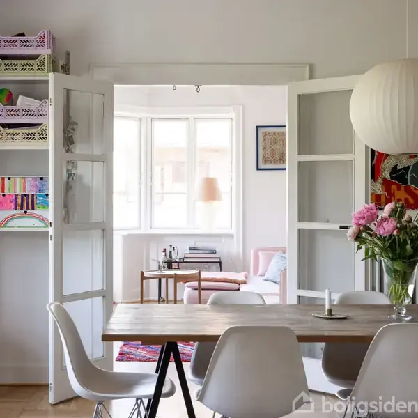 Et spisebord står i en lys stue. En døråbning fører til en opholdsstue med en sofa, et sidebord og vinduer. Lyserøde blomster pryder bordet. En lampe hænger fra loftet.