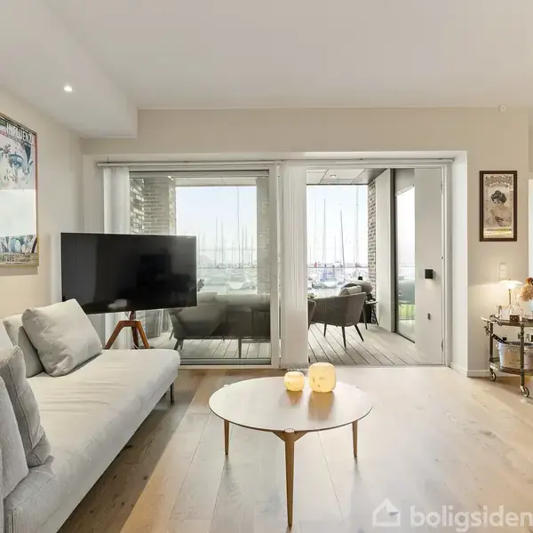 Stue med grå sofa og rund træbord, stor fladskærms-TV mod væg med kunstbilleder; store glasskydedøre åbner til balkon med udsigt til en havn.