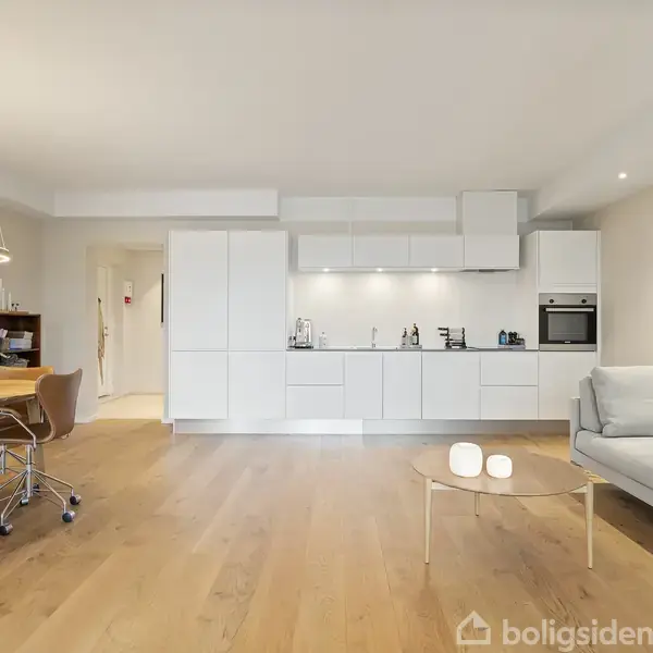 Lyst køkkenalrum domineret af hvide køkkenskabe strækker sig langs væggen. I forgrunden en sofa ved et rundt sofabord. Til venstre står et spisebord med stole.