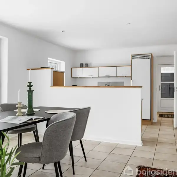 Spisebord med fire stole står i et lyst køkkenalrum. Bordet er dækket med stearinlys og dekorationer. I baggrunden ses et åbent køkken og en dør mod en entré.