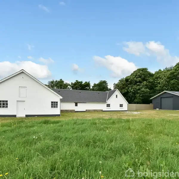 Hvid bygning med mørkt tag står i en åben mark. I baggrunden er en grå bygning og en række træer under en blå himmel med skyer.