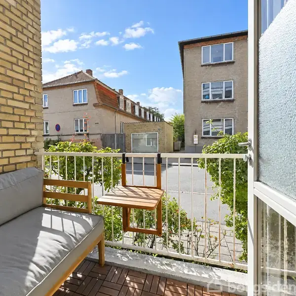 En lille balkon med en sofa og et lille bord, som vender ud mod en rolig gade med boligbygninger og grønne buske i forgrunden under en klar blå himmel.