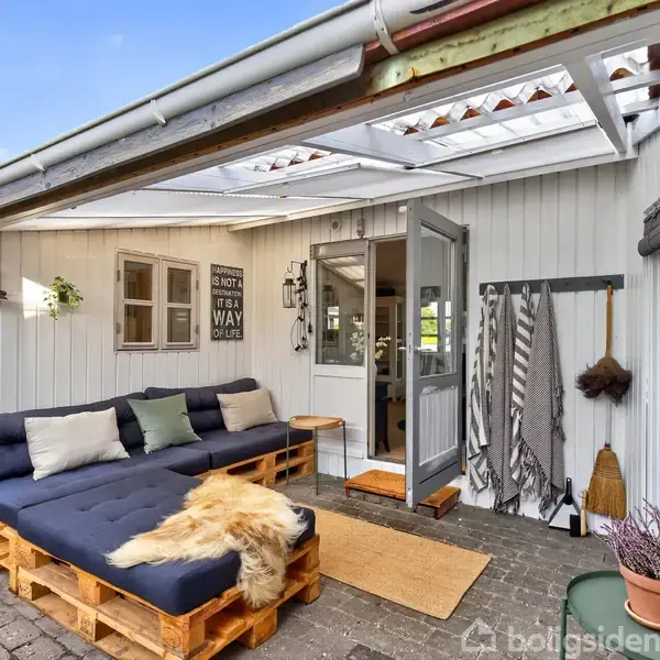 Udendørs loungeområde med blå pallesofa dekoreret med puder og pels. Dobbelt dør åben ind til huset. Tekst på skilt: "NOT A TINY HOUSE IT'S A WAY OF LIFE." Omkranset af lyse vægge og dekorationer.