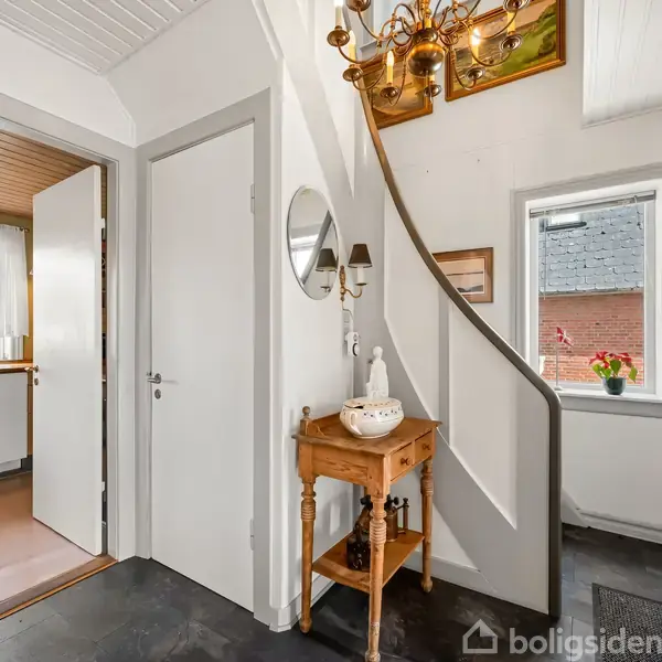 Rummelig entre med et lille træbord og spejl, oplyst af en lysekrone. En buet trappe fører opad. En åben dør viser en lys køkken med et vindue til haven.