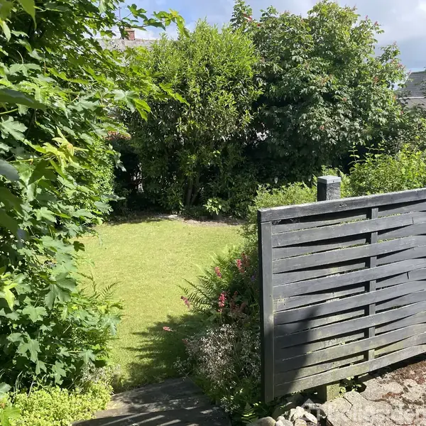Entré med træbord under en vægmonteret spejl og lampe. En trappe fører opad. Døråbning til køkken på venstre side med lysindfald fra vindue.