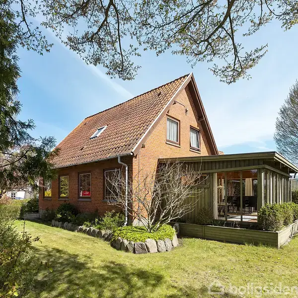 Murstenshus med rødt tag står i en have med græs og træer. En overdækket veranda er fastgjort til siden. Blåt himmelloft uden tekst i nærheden.
