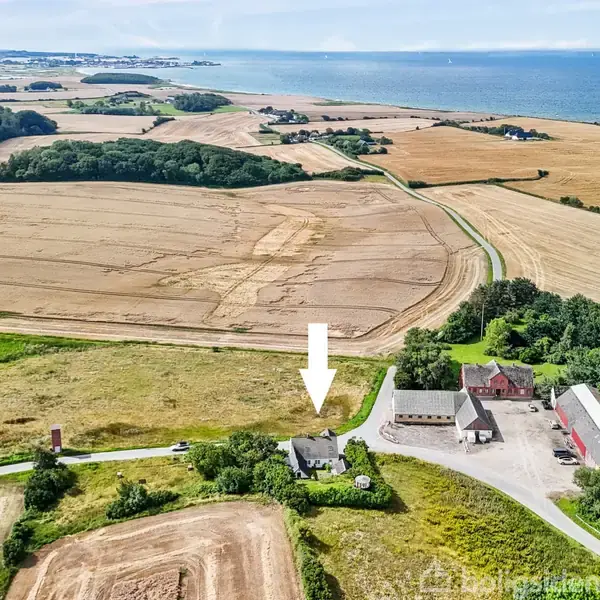 En hvid pil peger på et hus omgivet af marker og grusveje. I baggrunden ses et kystlandskab med havet og spredte bygninger. Markerne er delvist høstede.