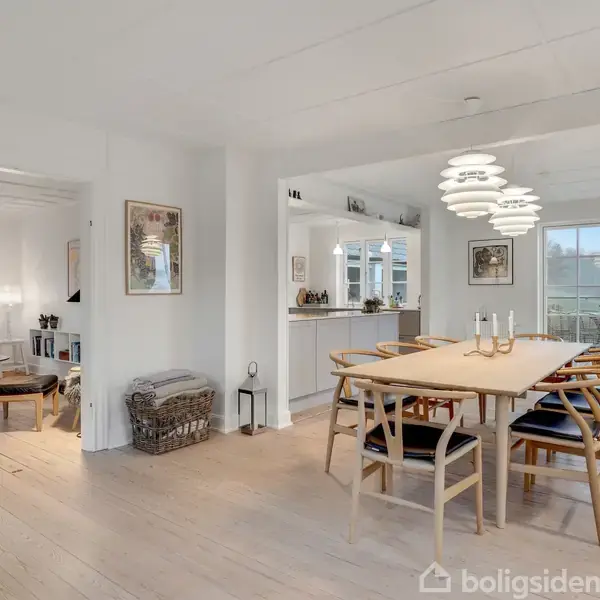 Et spisebord omgivet af otte stole står i en lys spisestue. En moderne lampe hænger over bordet. I baggrunden ses en stue og en åben køkkenafdeling.