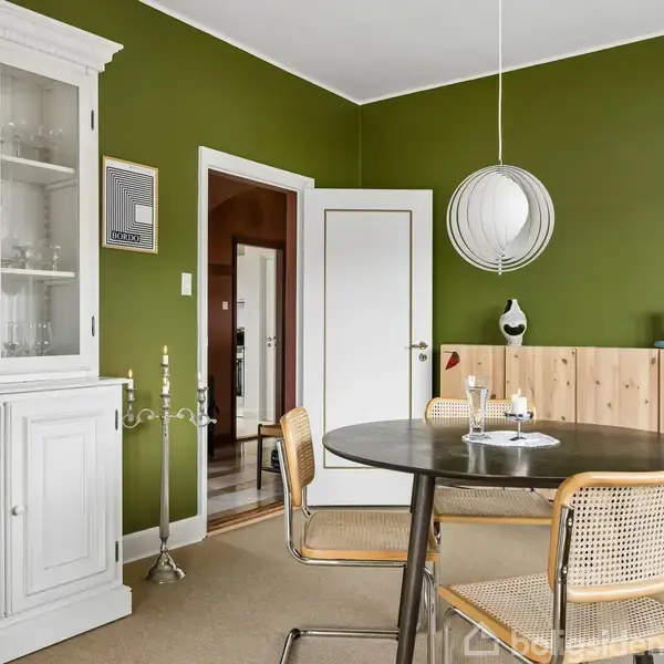 Et rundt spisebord med stole står i et rum med grønne vægge. Et hvidt vitrineskab står til venstre, og en dekorativ lampe hænger over bordet. Ingen tekst er synlig.