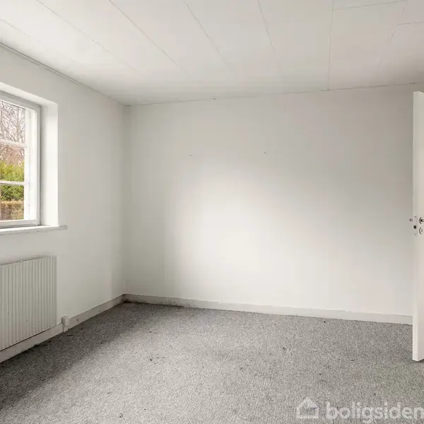 Et tomt værelse har et åbent vindue mod venstre og en åben dør mod højre. Væggene er hvide, gulvet er dækket af gråt tæppe.