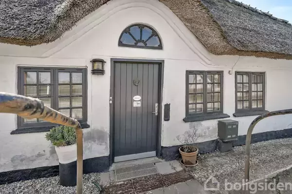 En dør med sort ramme og buet vindue, flankeret af to vinduer i en hvidkalket mur. Et stråtækt tag over og grus med planter i potter foran.