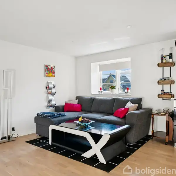 En grå hjørnesofa med røde puder står i et moderne, lyst rum. Et sofabord med hvidt stel står på et sort tæppe. Ved siden af ses en hylde og et lille barbord. En vægudsmykning hænger over sofaen.