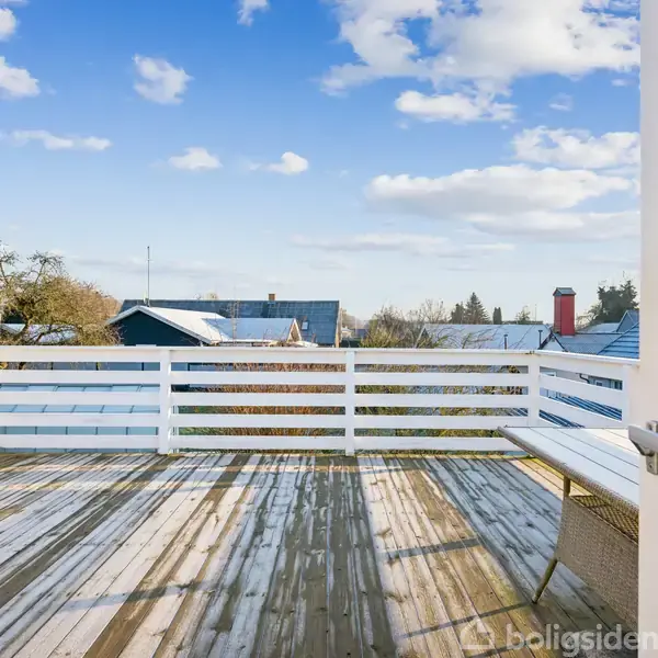 En åben træterrasse med hvidt rækværk, delvist dækket af let sne. Terrasse udsigt over sneklædte hustage og træer, klar himmel i baggrunden. Ingen tekst fundet.