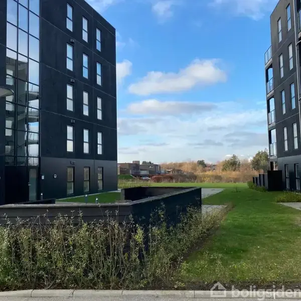 Bygninger står stille overfor hinanden i et beboelsesområde. En grøn have med græsplæner strækker sig mellem dem. Blå himmel og lette skyer kan ses i baggrunden.