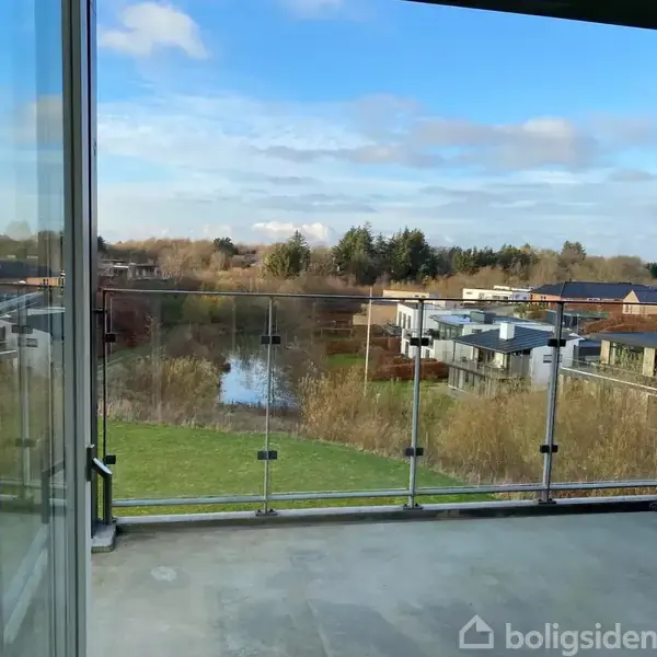 Balkon med glasrækværk kigger ud over en grøn park med en lille sø. I baggrunden ses moderne huse og træer under en delvist skyet himmel.