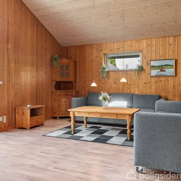 Gråt hjørnesofa sætter ved et træbord i en stue med trævægge og loft. Lamper hænger ned over bordet. Store vinduer giver adgang til en terrasse.