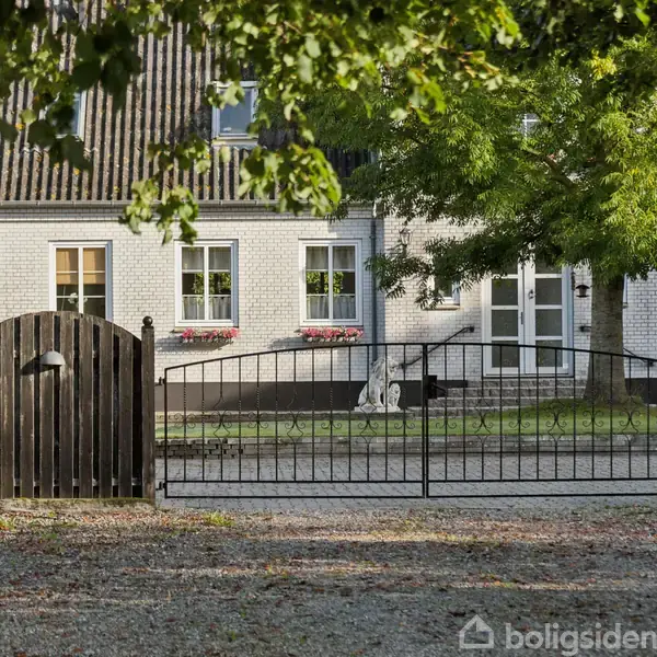 Hvid murstensbygning med sort tag står bag en sort smedejernsport. Træer omkranser området, og grus dækker jorden foran. Vinduernes blomsterkasser tilføjer farve til facaden.