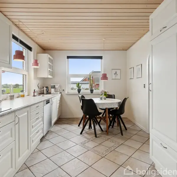 Et lyst køkken med hvidt inventar og klinkegulv. Et rundt bord med fire stole står midt i rummet. Vinduerne giver udsigt til grønne områder udenfor.