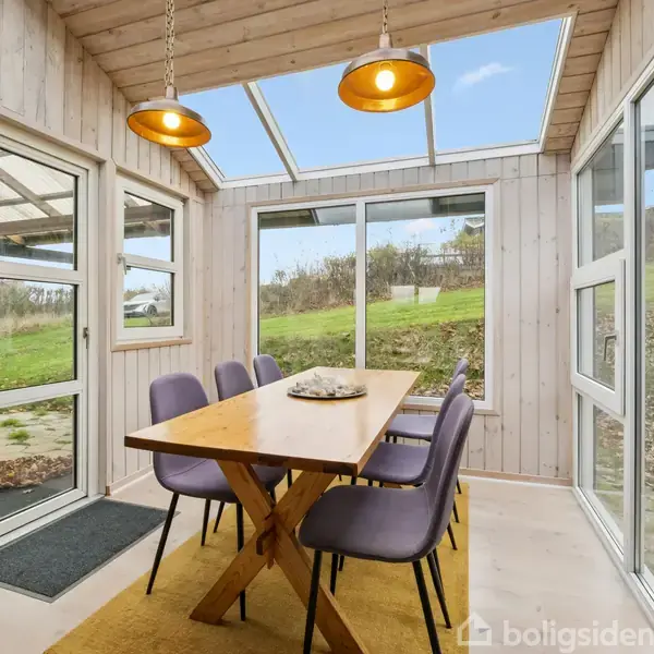Et træspisebord med lilla stole står i et lyst udestueområde med store vinduer. Det er udsigt til en græsplæne udenfor. To gule lamper hænger over bordet.