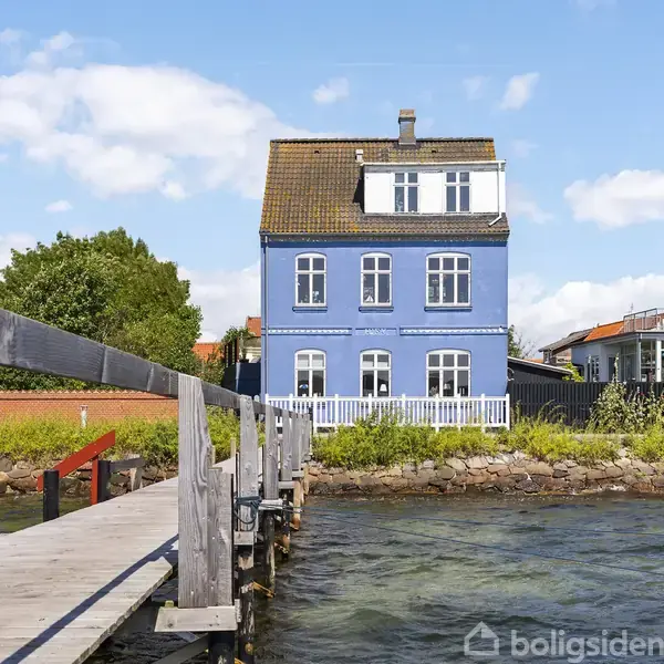 Blåt hus står ved en kaj med træbro, omgivet af vand og en grøn have. En anden bygning ses til højre under en klar, blå himmel.