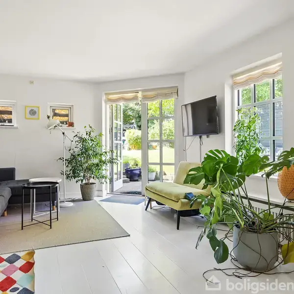 Stue med sofa og grøn stol, omgivet af planter. Et tv hænger på væggen. Store vinduer og en dør åbner ud til en frodig have. Farverigt tæppe på gulvet.