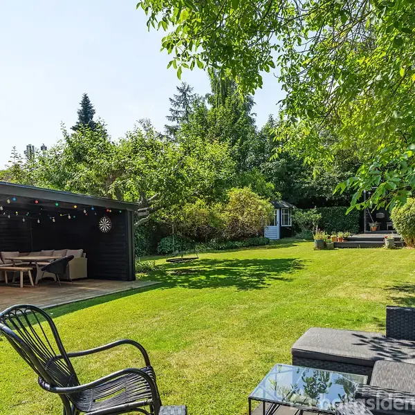 En have med en overdækket terrasse indeholder spisebord og stole. Græsplænen er omgivet af buske og træer. Et loungeområde med sofa og bord ses i forgrunden under træer.