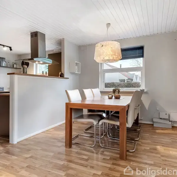 Spisebord med fire stole står i en lys spisestue med trægulv. En pendel hænger over bordet. Til venstre ses et køkken med halvåben væg og hylder.