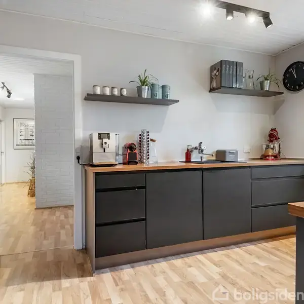 Køkken med mørke skabe og træbordplade. Kaffemaskine og dekorationer står ovenpå. Væg med hylder, ur til højre. Trægulv og åben døråbning til lys gangområde.