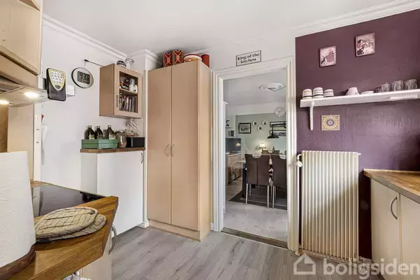 Et køkken med træmøbler og køkkenudstyr på bordet, set mod en spisestue. En hylde med glas og dekorationer ses på en lilla væg. Skiltet lyder "Home of the cat kitchen".