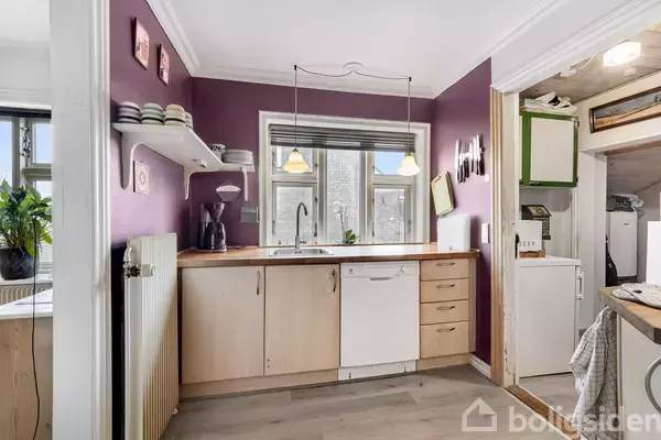 Køkken med træskabe og en opvaskemaskine under en bordplade, to hængende lamper over vasken, omgivet af lilla vægge. Der er hylder med tallerkener, og et tilstødende bryggers er synligt.