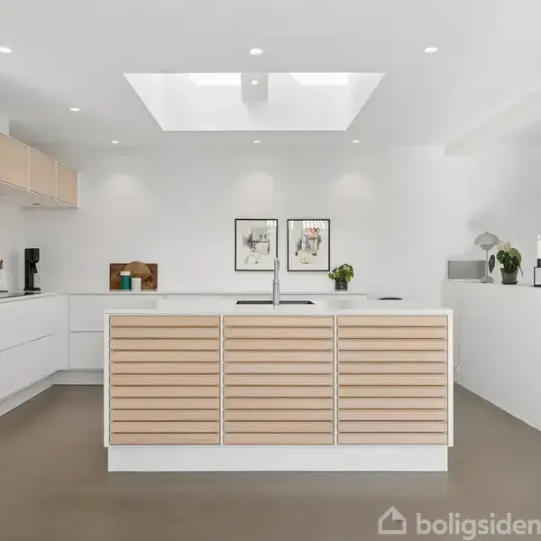 Et moderne køkken med en central ø med trælamel-front, omgivet af hvide skabe og apparater. Loftvinduet lader lys ind, mens vægge prydes med kunstværker og grønne planter.