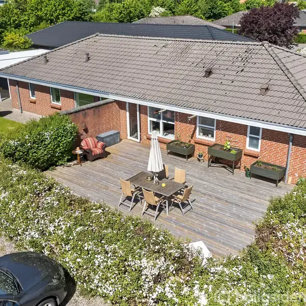 En etplans murstenshus med gråt tag har en stor træterrasse foran. Terrassen er udstyret med et bord, stole og en parasol. Omkring ligger en velplejet have.