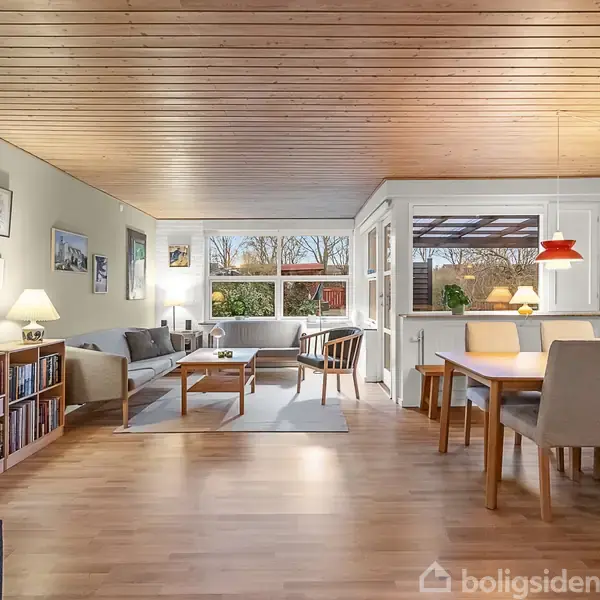 Stuen indeholder en lysegrå sofa, sofabord og reol med bøger. Store vinduer giver udsigt til en have. En spisestue med træbord og stole er til højre for sofaområdet.
