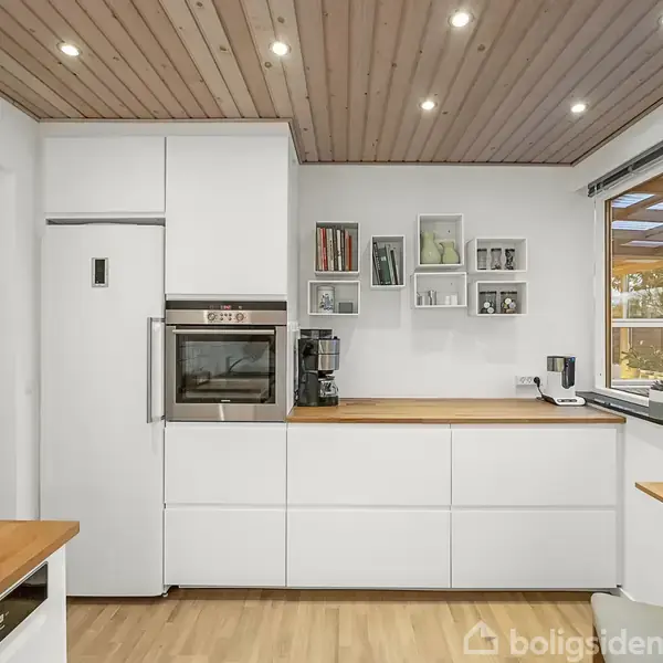 Et lyst køkken med hvide skabe, en indbygget ovn og en kaffemaskine på bordpladen. Der er hylder med bøger og billeder over bordet. Vindue med udsigt til terrasse.