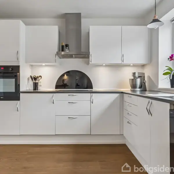 Et moderne, funktionelt køkken med hvide skabe, grå bordplader og indbygget ovn. En vase med blomster står ved håndvasken under et stort vindue, som giver udsigt til et boligområde.