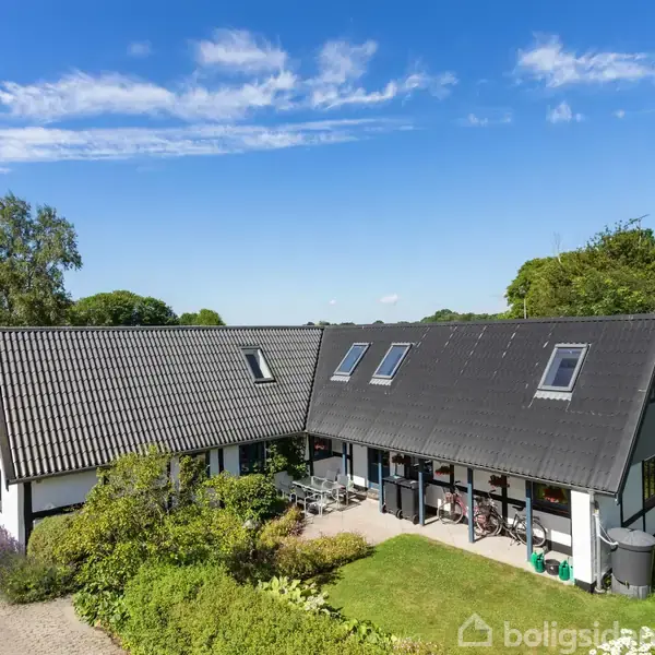 Et hvidt hus med sort tag står i en grøn have. En terrasse med møbler er foran huset. Blå himmel med få skyer i baggrunden.