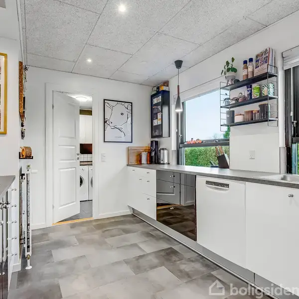 Et moderne køkken med hvidt inventar strækker sig i vinkler, og store vinduer lukker lys ind. Væggene har kunstværker, og hylder rummer flere objekter. En åben dør fører til et tilstødende rum.