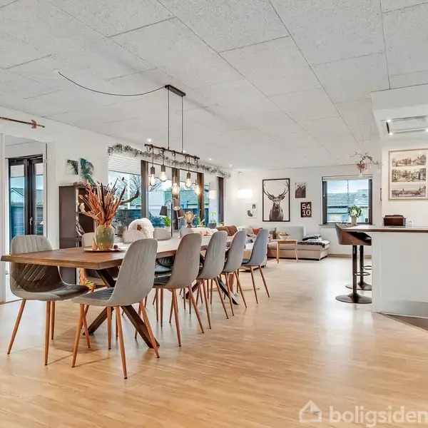 Stue med grå hjørnesofa foran store vinduer, dekoreret med puder. Vægdekoration inkluderer et hjorteprint og "54 10" billede. Fjernsyn og møbler står ved en væg.